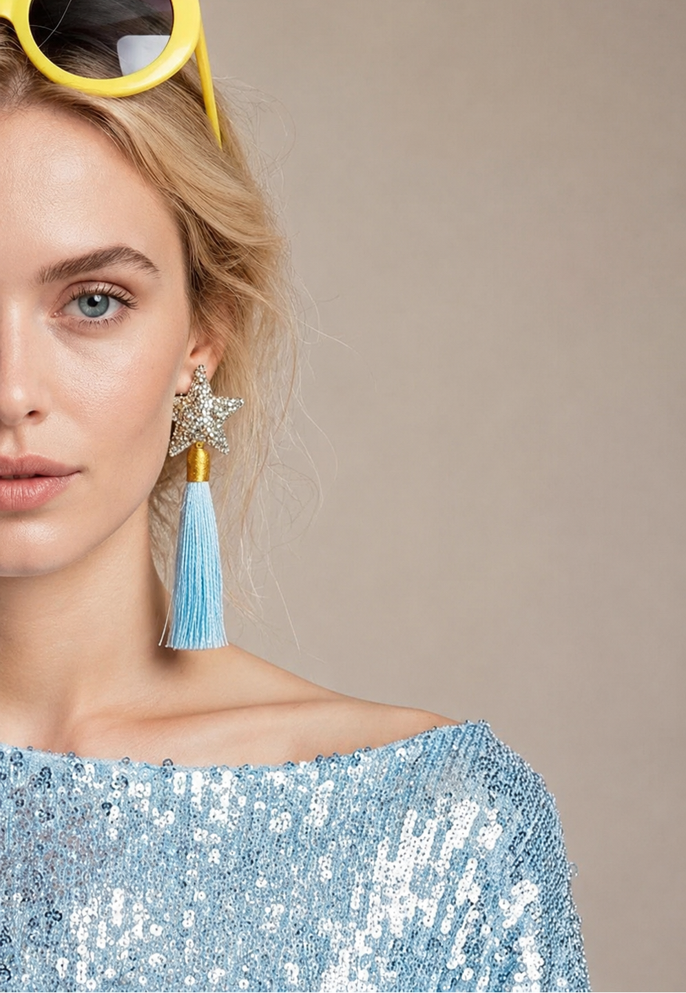A close-up of a woman wearing light blue tassel earrings with star-shaped studs and a light blue sequined top, with yellow sunglasses on her head against a neutral beige background.